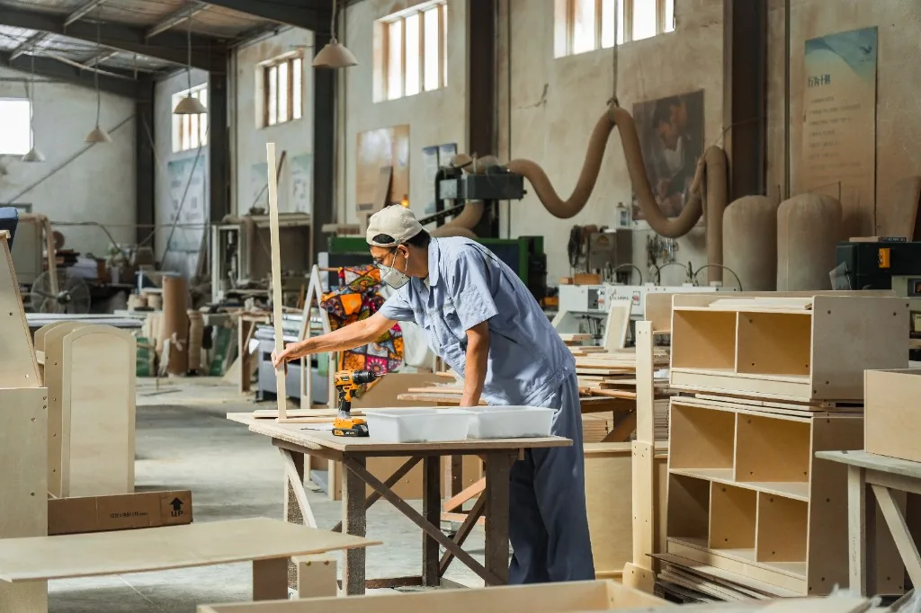 Factory worker in a blue coverall assembling a birch plywood preschool cubby at a bench